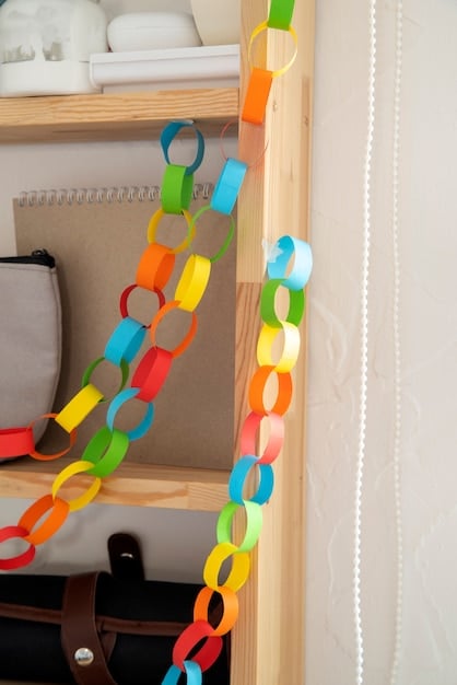 A close-up of a handcrafting paper garland. The garland is made of colorful, patterned paper cut into various shapes like stars and circles. The hand is carefully threading the shapes together using a needle and thread. The background is a workspace with various craft supplies.