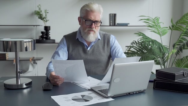 A senior man sitting at a desk, carefully using a laptop, looking at a budgeting spreadsheet with various financial figures, symbolizing organized debt management.